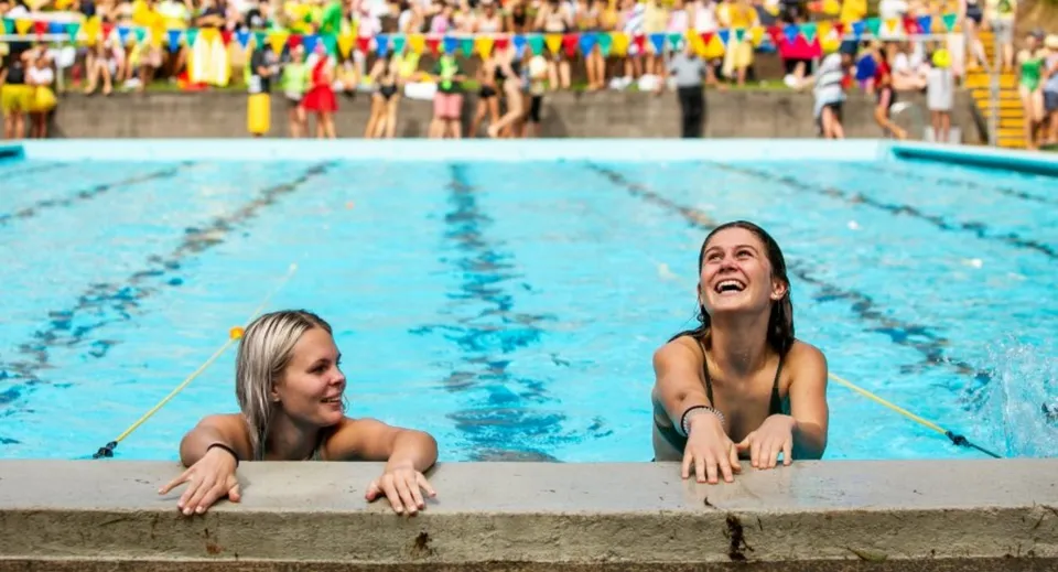 Due donne siedono nella piscina nell'area del Tauranga Girl´s College, mentre sullo sfondo si vedono una folla di persone e bandiere colorate.