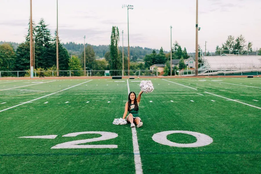 Una animadora de Redmond High School está de pie en el campo de césped artificial frente a una zona boscosa y edificios.