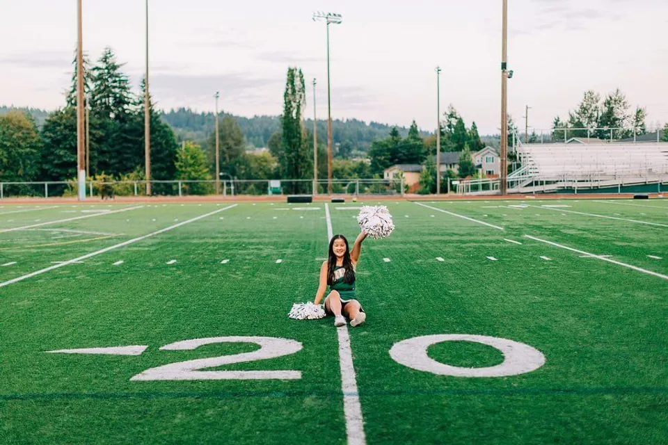 Una cheerleader della Redmond High School si trova sul campo in erba sintetica davanti a un'area boschiva ed edifici.