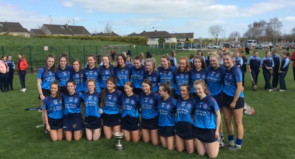 Un gruppo di giovani atlete della Meán Scoil Mhuire Longford posa con un trofeo su un campo in erba davanti a delle abitazioni.