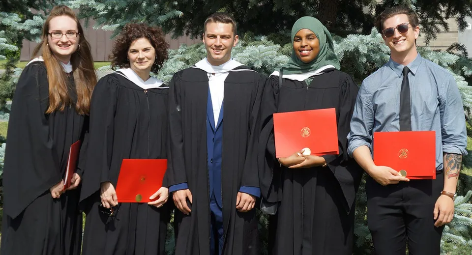Cinque persone in abiti accademici sono in piedi nel campus della South Carleton High School davanti ad alberi e aree verdi.