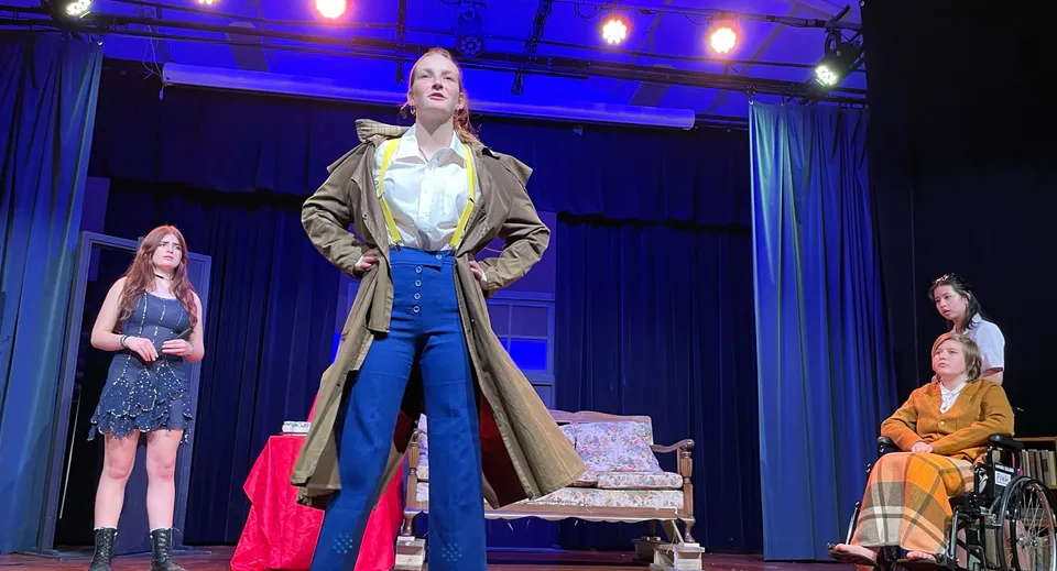 Una donna con un lungo cappotto beige è sul palco del Waiuku College, circondata da altre donne in abiti colorati.