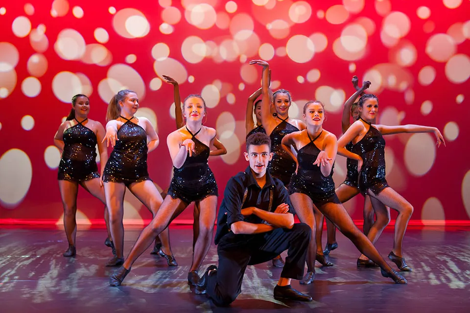 Un gruppo di ballerini e ballerine della Davidson High School si esibisce su un palco davanti a uno sfondo rosso a pois bianchi.