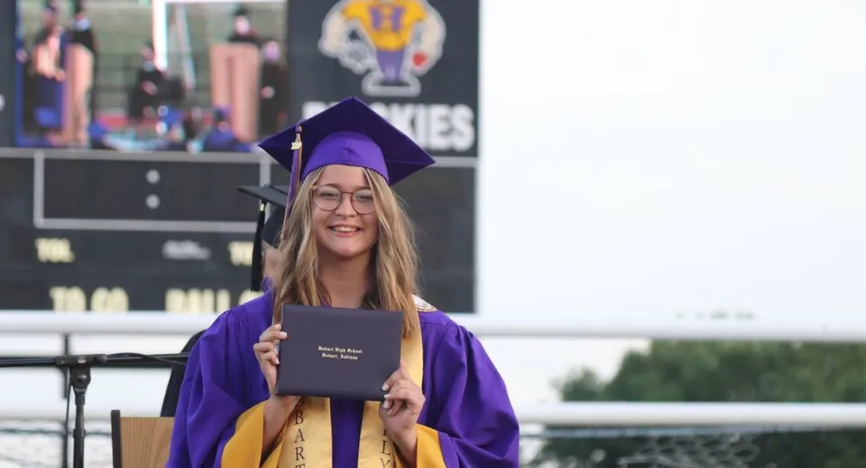 Una studentessa della Hobart City High School indossa una toga viola da diploma e tiene in mano il suo diploma davanti a uno sfondo con il logo dell'università.