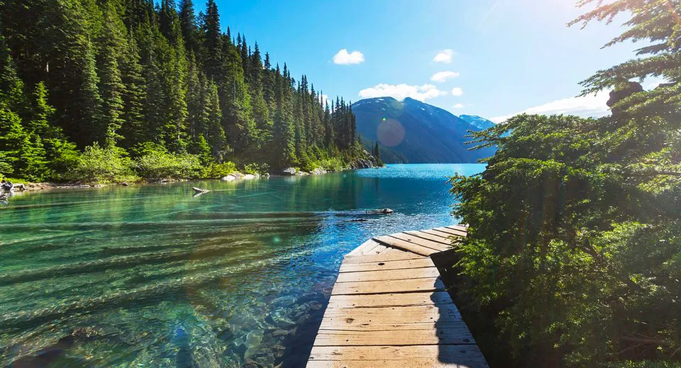 Un pontile in legno si estende su un lago limpido vicino alla South Delta Secondary School con montagne e foreste sullo sfondo.