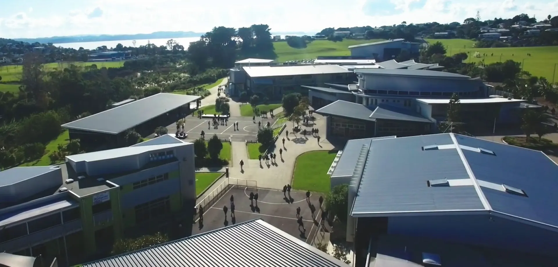 Il campus del Whangaparāoa College con edifici moderni, sentieri e spazi verdi davanti a un paesaggio collinare e uno specchio d'acqua.