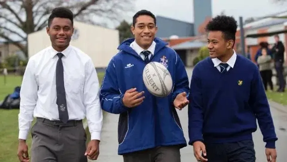 Tre studenti della Timaru Boys School in divisa scolastica con un pallone da rugby davanti a un edificio scolastico e ad alberi.