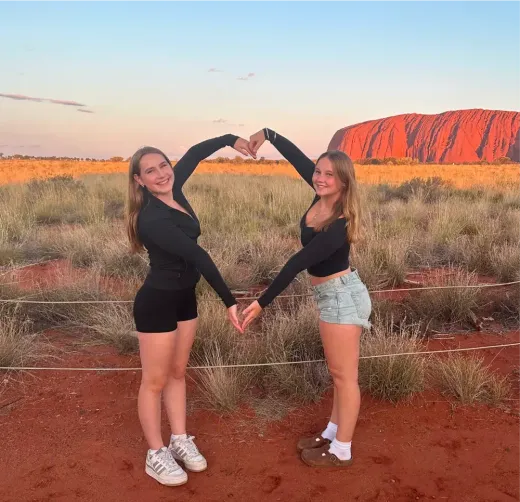 Zwei Mädchen formen bei Sonnenuntergang vor dem Uluru ein Herz mit den Armen in Australien