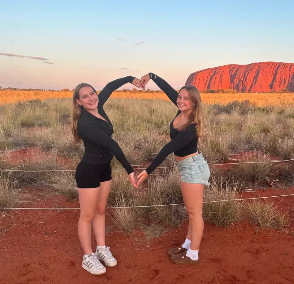 Zwei Mädchen formen bei Sonnenuntergang vor dem Uluru ein Herz mit den Armen in Australien