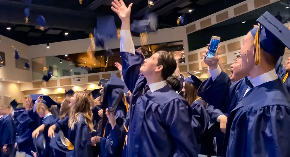 Un gruppo di diplomati della Colorado Springs Christian School festeggia in toga blu e con le braccia alzate in un ambiente interno festosamente illuminato.