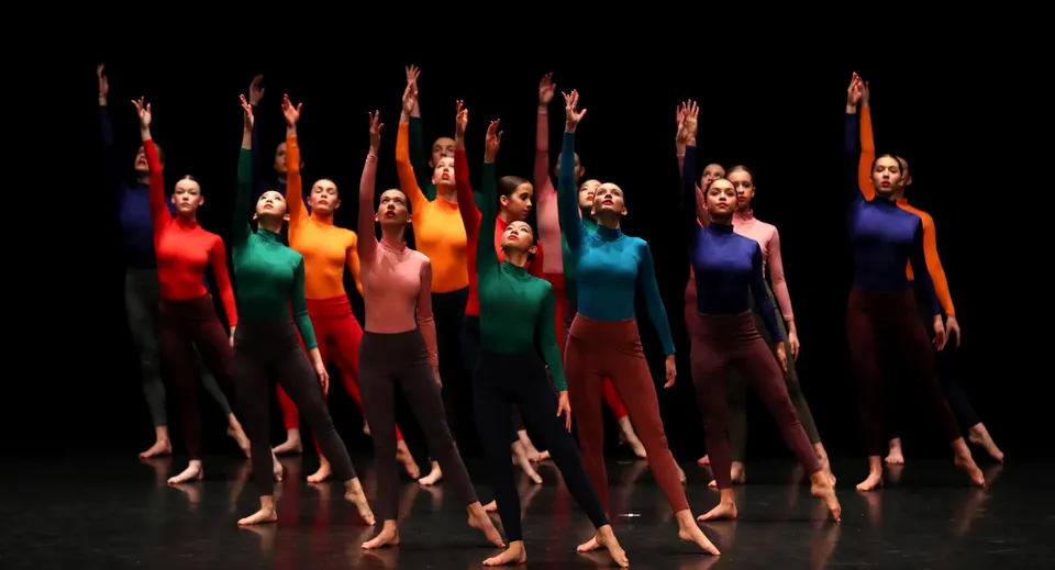 Un gruppo di ballerine della St. Edmunds School esegue su un palco buio una coreografia sincronizzata con le braccia alzate.