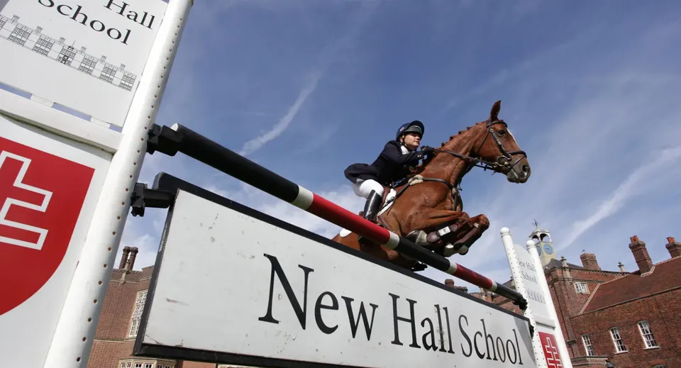 Una studentessa della New Hall School a cavallo mentre salta un ostacolo davanti a un edificio scolastico.