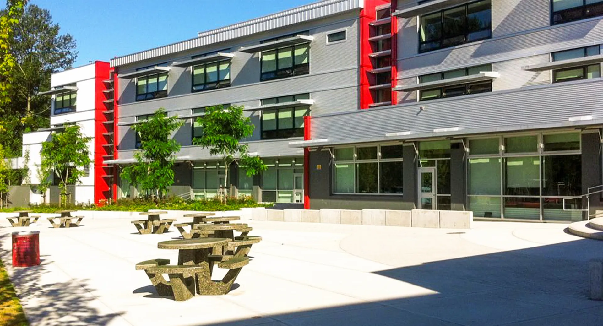 L'edificio moderno a più piani della Carson Graham Secondary School con grandi finestre e dettagli rossi si trova dietro un cortile pavimentato con panchine in legno.