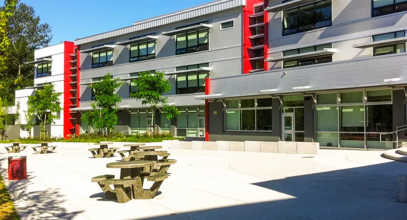 L'edificio moderno a più piani della Carson Graham Secondary School con grandi finestre e dettagli rossi si trova dietro un cortile pavimentato con panchine in legno.