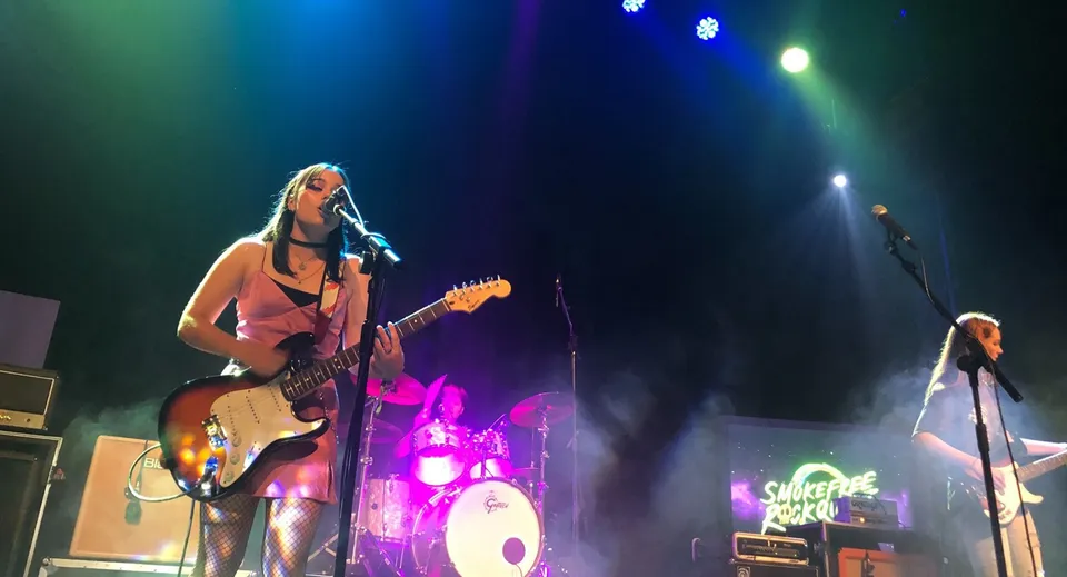 Una studentessa della New Plymouth Girls’ High School suona la chitarra elettrica sul palco con la band e la batteria sullo sfondo.