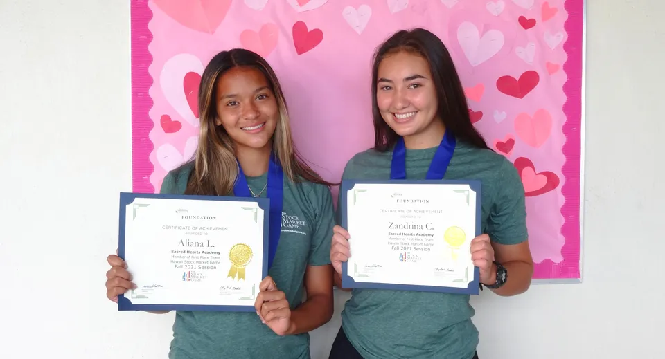 Due studentesse della Sacred Hearts Academy mostrano i loro certificati davanti a uno sfondo rosa con motivi a cuore.
