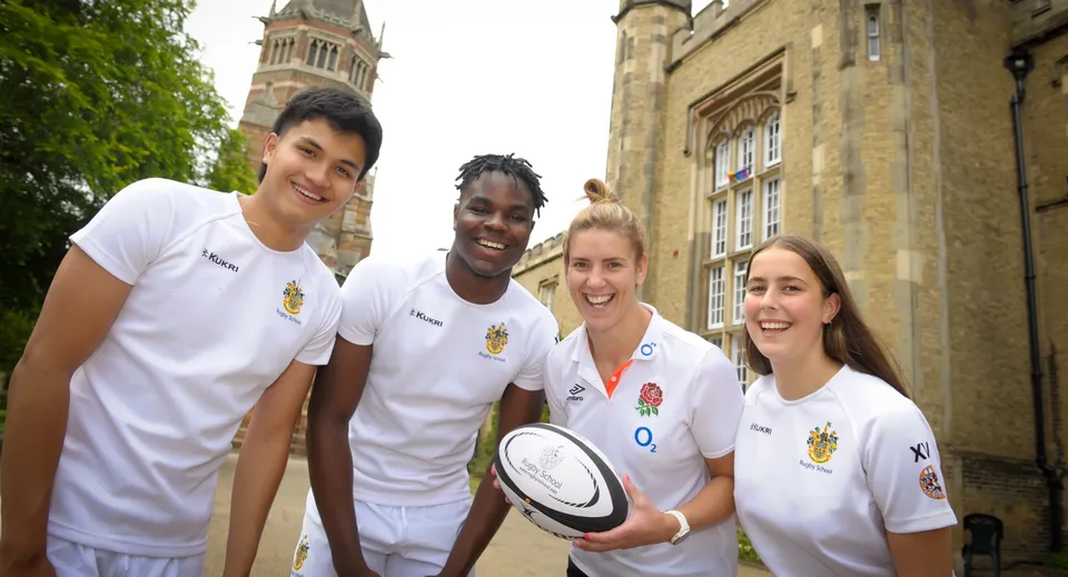 Quattro studenti della Rugby School si trovano davanti a un edificio storico con un'alta torre, circondati da una verde vegetazione.