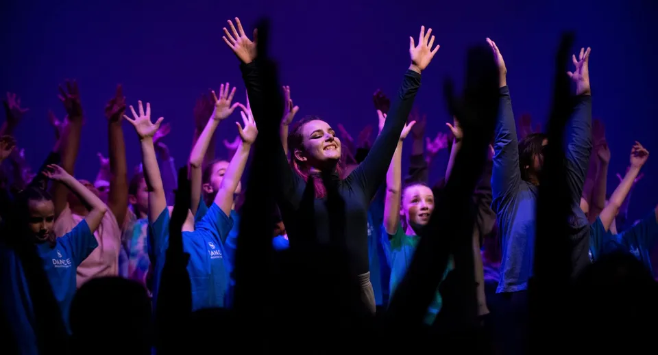 Un gruppo di studentesse della Brighton Girls alza le braccia in segno di esultanza davanti a uno sfondo scuro con illuminazione viola-blu.