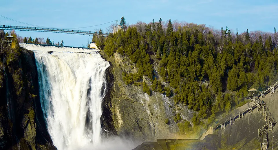 Una cascata scende lungo un pendio roccioso davanti a un bosco e a un ponte sospeso nel campus della Windermere Secondary School.