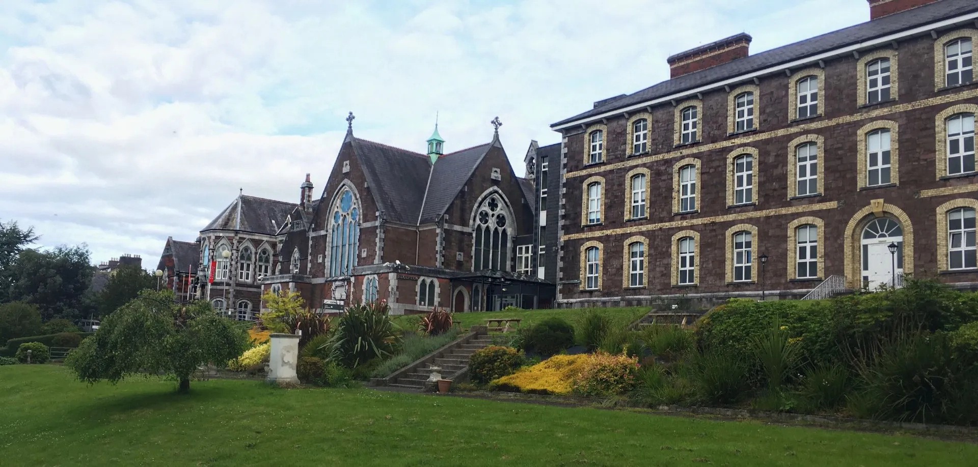 L'edificio storico della Cork Educate Together Secondary School con un giardino ben curato in primo piano sotto un cielo nuvoloso.