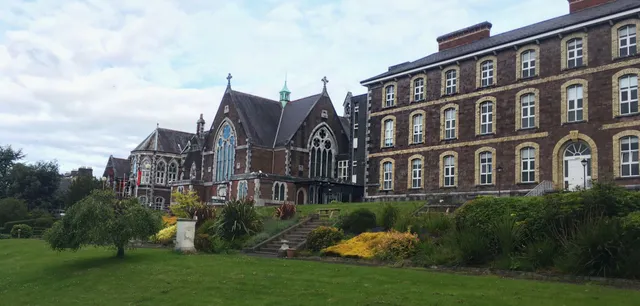 L'edificio storico della Cork Educate Together Secondary School con un giardino ben curato in primo piano sotto un cielo nuvoloso.