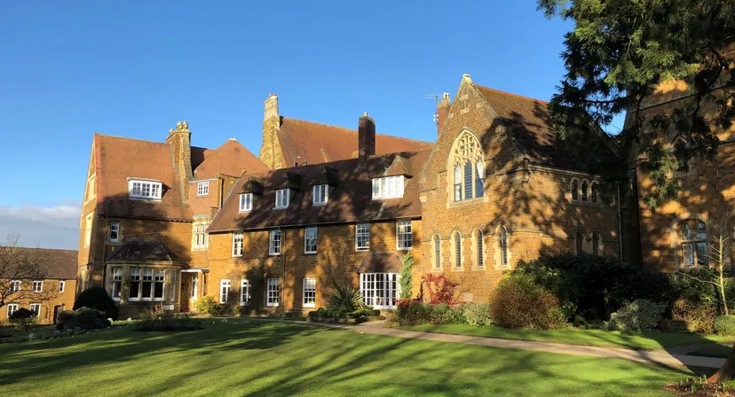 L'edificio storico in mattoni della Bloxham School sorge circondato da un prato curato e aree verdi sotto un cielo limpido.