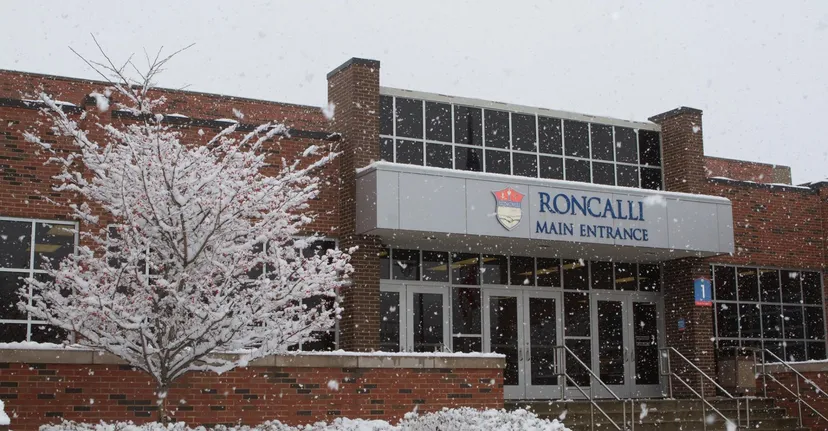Un albero coperto di neve si trova davanti all'edificio in mattoni con l'ingresso principale della Roncalli Central High.