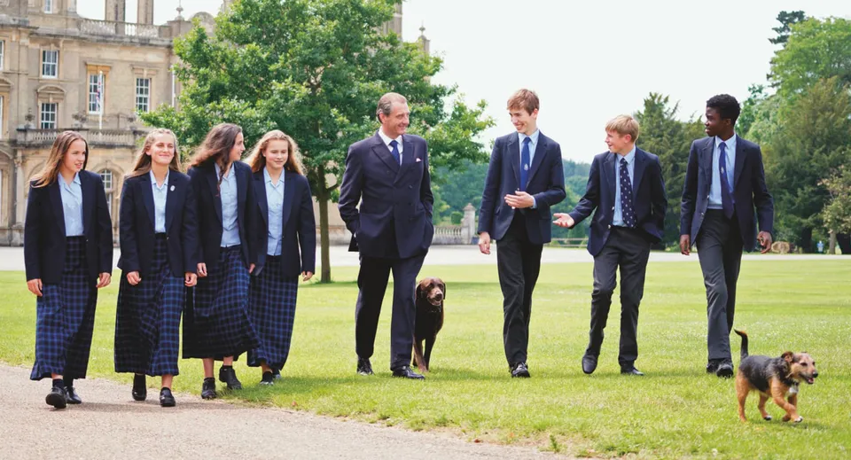 Un gruppo di studenti della Culford School cammina insieme a un cane su un prato tra alberi ed edifici.