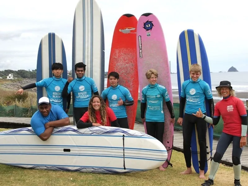 Estudiantes de la New Plymouth Boys' High School posan con tablas de surf frente a un paisaje costero con montañas al fondo.