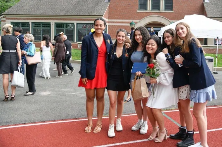 Seis jóvenes sonrientes posan juntas en el exterior de un colegio sosteniendo un diploma y flores