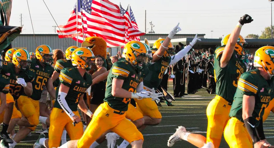 Un gruppo di giocatori di football della Bishop Carroll High School si trova sul campo da gioco davanti a uno stadio con bandiere americane.