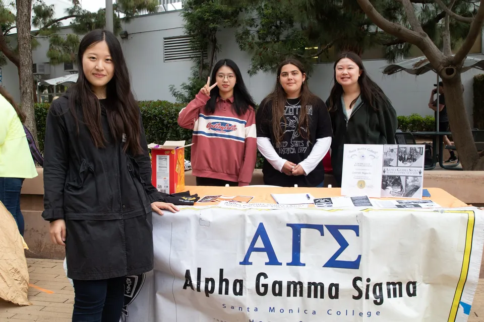 Quattro studentesse stanno dietro un tavolo con uno striscione di Alpha Gamma Sigma sul campus del Santa Monica College.