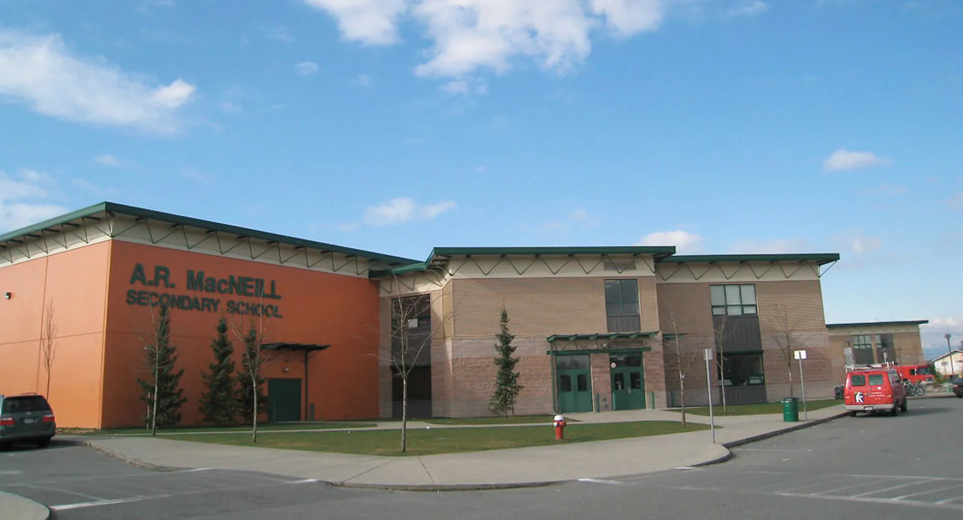 L'edificio scolastico moderno a più piani della A.R. MacNeill Secondary School con facciata rossa e un ingresso in evidenza sotto un cielo blu.