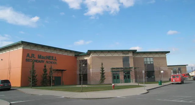 L'edificio scolastico moderno a più piani della A.R. MacNeill Secondary School con facciata rossa e un ingresso in evidenza sotto un cielo blu.