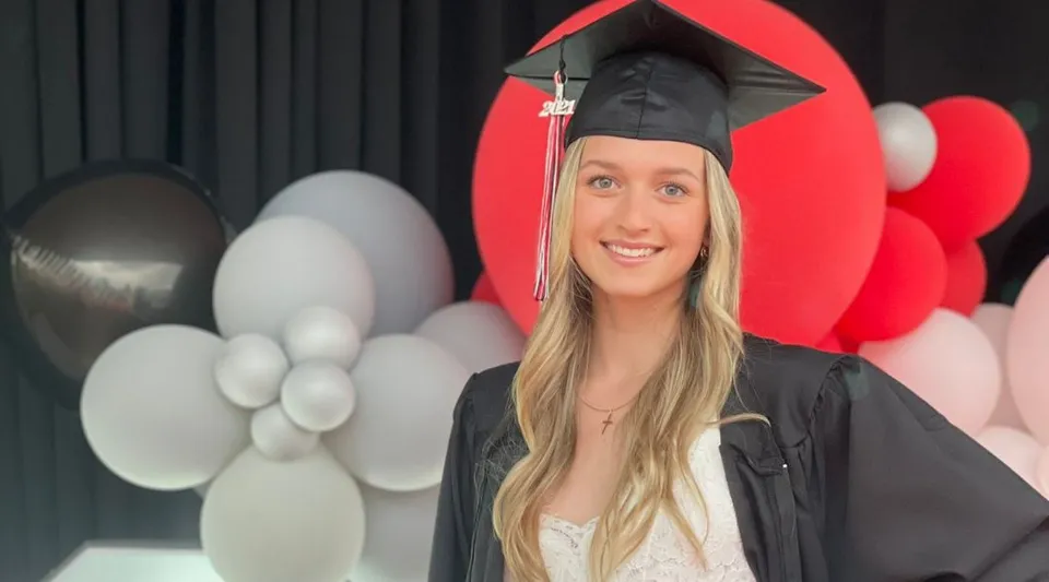 Una studentessa della Maxwell Heights Secondary School in toga da diploma sta davanti a palloncini rossi e bianchi.