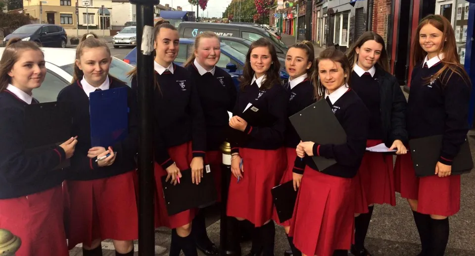 Studentesse del St. Dominic’s College in uniforme scolastica si trovano in strada con edifici e veicoli sullo sfondo.