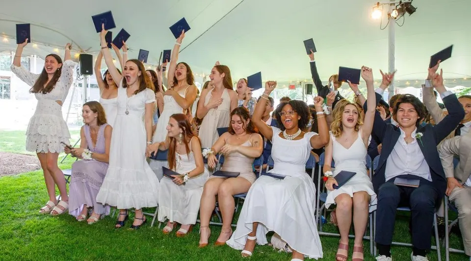 Un gruppo di studentesse della Cambridge School of Weston festeggia all'aperto la propria cerimonia di diploma con abiti bianchi e cappelli da laurea.