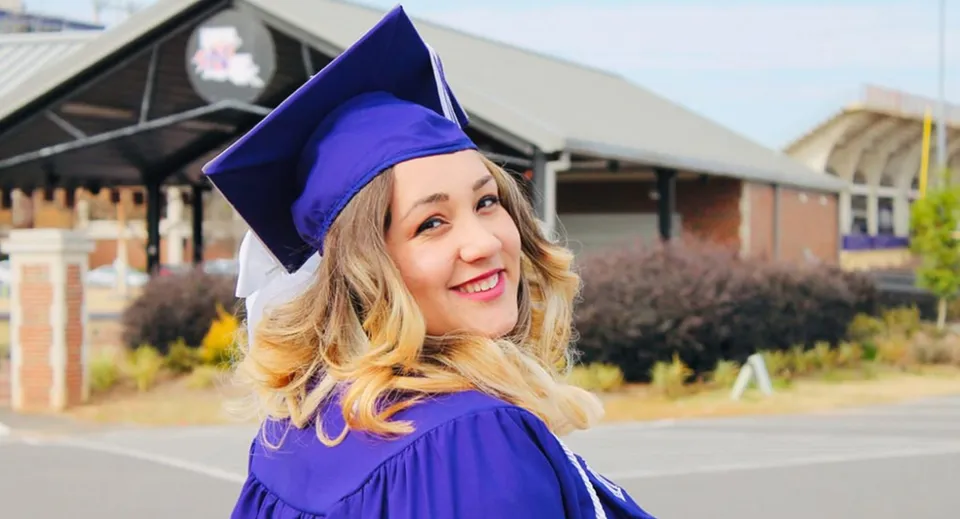 Una giovane donna sorridente con la toga di diploma viola è in piedi davanti a un edificio del campus della Sir Wilfrid Laurier Secondary School.