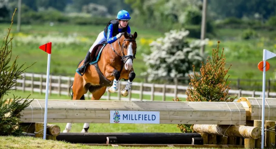Una studentessa della Millfield School va a cavallo con casco e abbigliamento protettivo, saltando un ostacolo di legno nel verde.