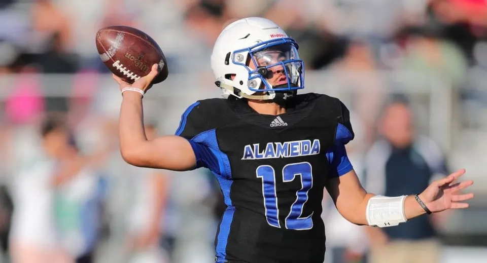 Un giocatore di football americano in maglia nera e blu tiene la palla sul campo da gioco dell'Alameda Unified School District.