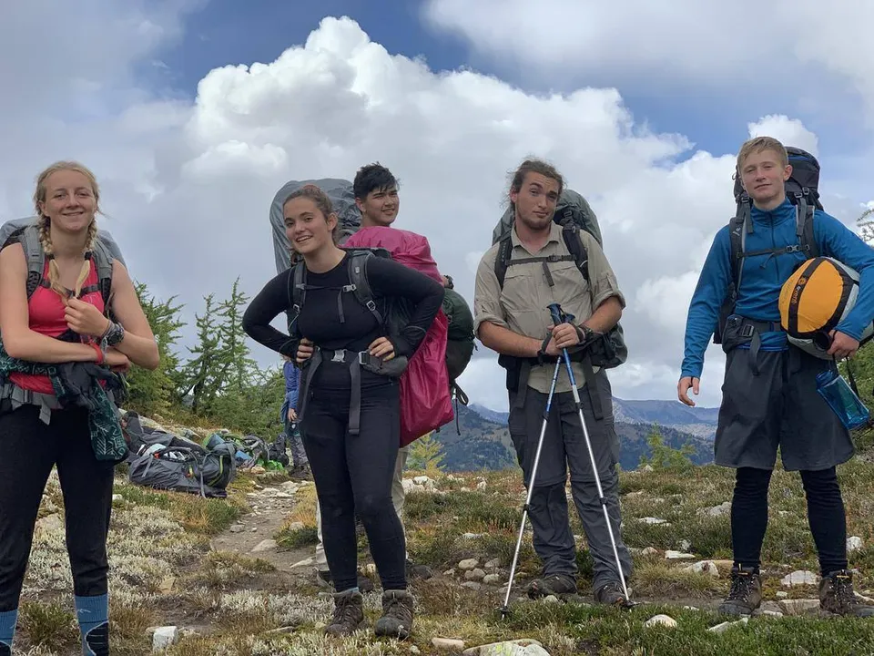 Un gruppo di studentesse della Spring Street International School in abbigliamento da trekking si trova in un paesaggio montuoso con nuvole e cielo blu.