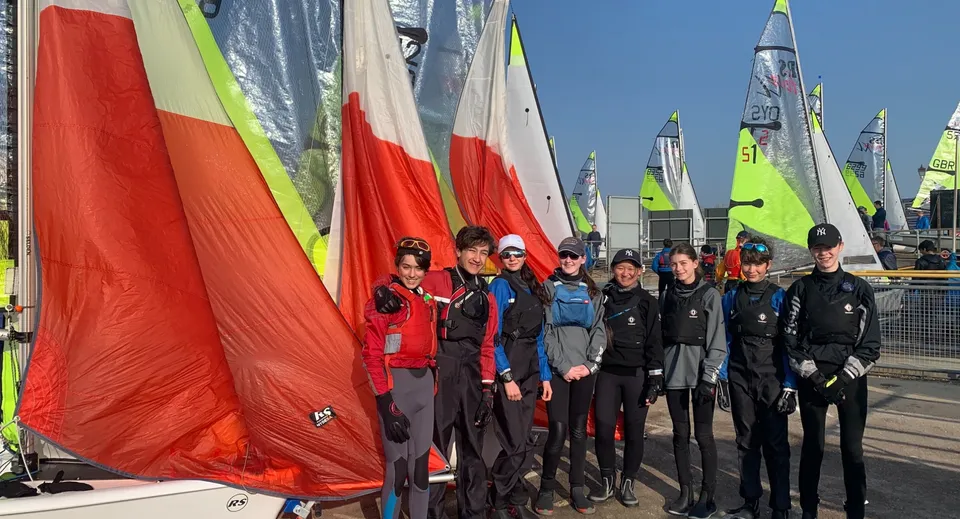 Un gruppo di studenti della Harrow School in abbigliamento da vela colorato sta davanti a una fila di barche a vela con vele rosse e verdi.