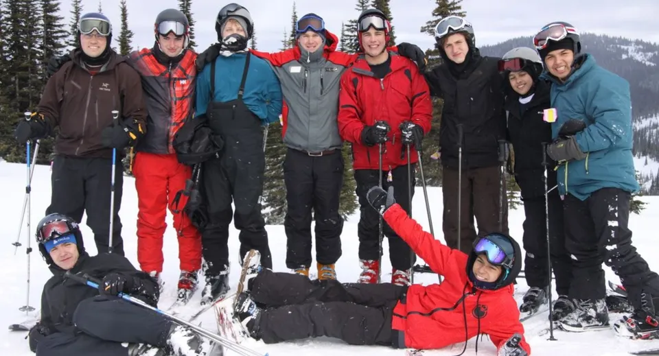Un gruppo di studenti della St. George’s School in abiti invernali colorati si trova su un pendio innevato con un bosco sullo sfondo.
