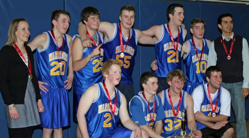 Un gruppo di giovani giocatori di basket della Athens District High School con maglie blu è davanti a una parete blu e tiene in mano delle medaglie.