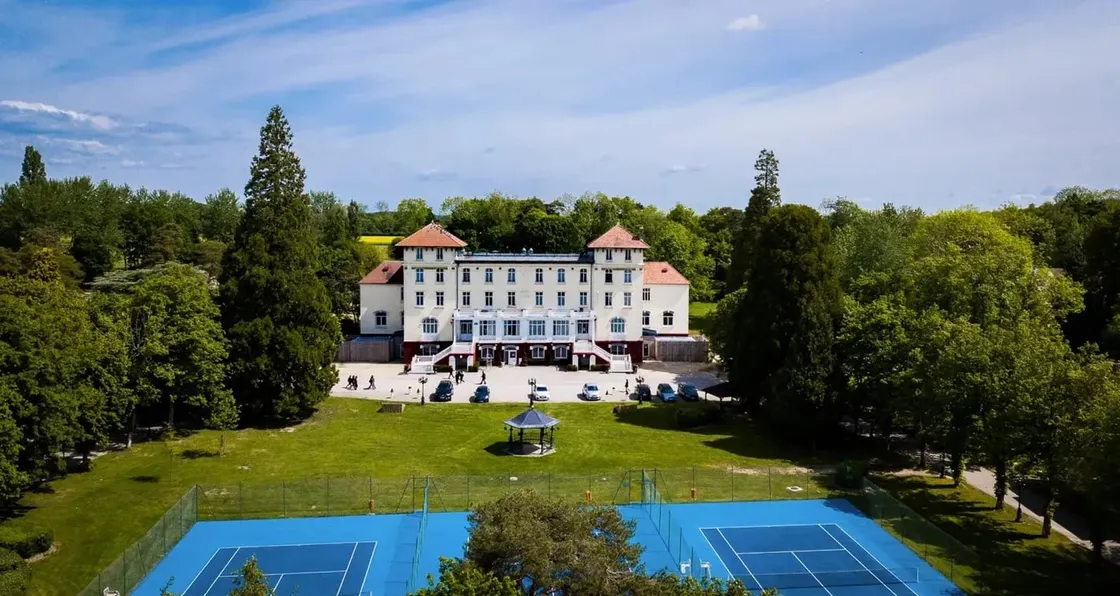 Luftaufnahme eines Internatsgebäudes mit Türmchen und rotem Ziegeldach, umgeben von Parkanlage und Tennisplätzen