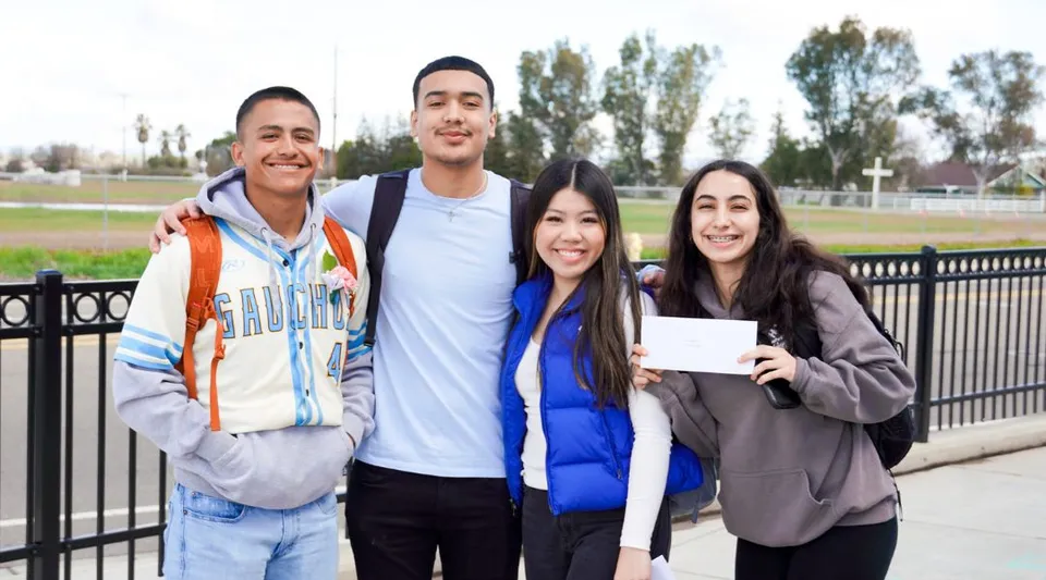 Quattro studenti del Merced Union High School District si trovano all'aperto in un'area simile a un parco con alberi e ringhiere.