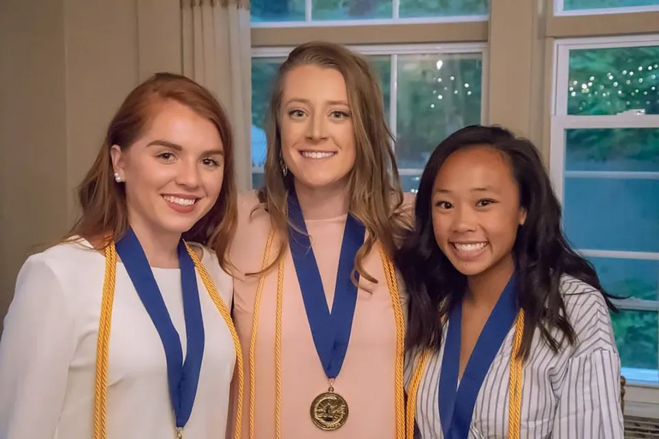 Tre laureate della Stearns High School in abito di laurea con medaglie davanti a una finestra con vista sul verde.