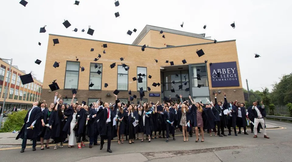 Un gruppo di diplomati dell'Abbey College Cambridge lancia in aria i propri tocchi davanti a un edificio in mattoni.