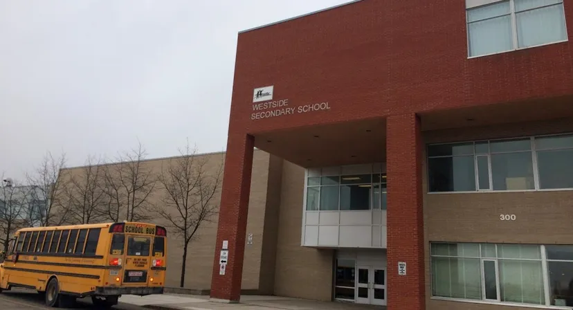Si vede l'edificio in mattoni della Westside Secondary School con uno scuolabus giallo davanti.