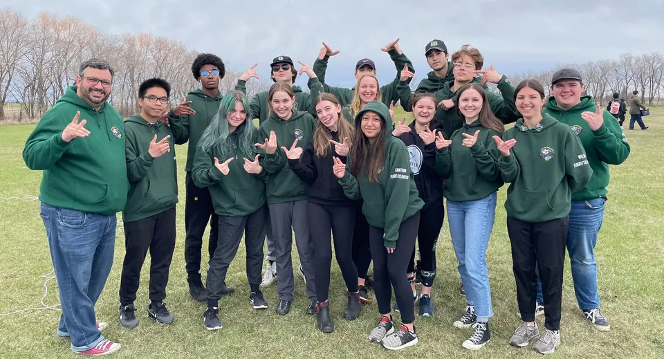 Un gruppo di studenti del Miles Macdonell Collegiate si trova in un campo all'aperto indossando felpe verdi con cappuccio.
